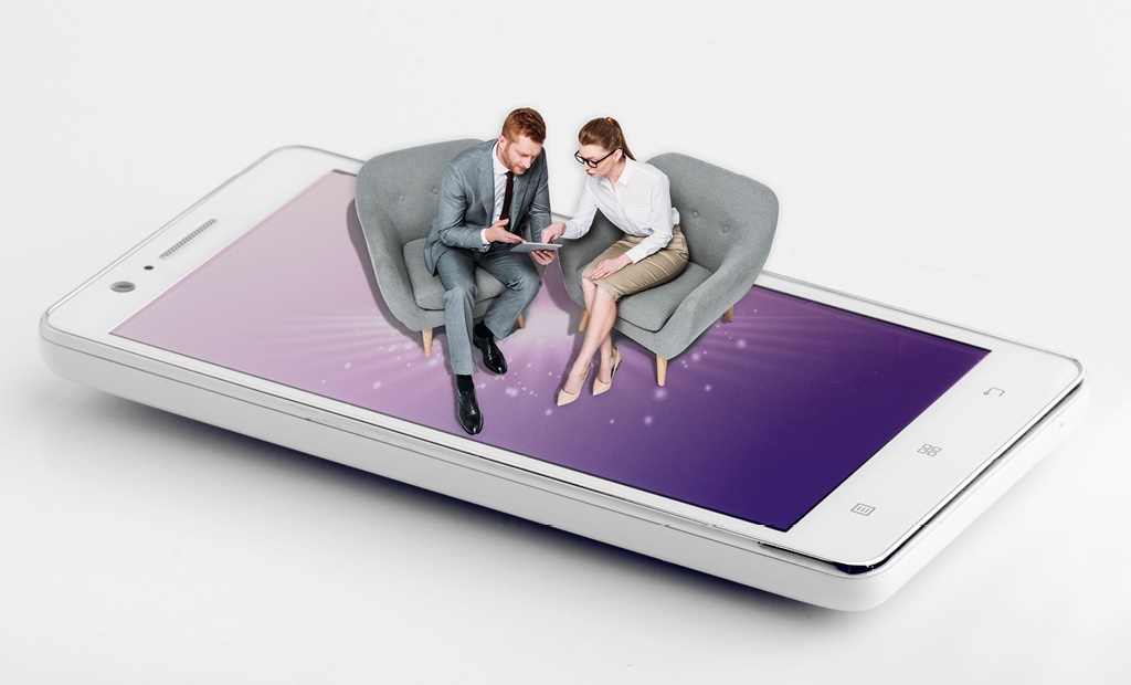 Two businesspeople sit on armchairs placed on top of a large mobile phone screen, discussing something on a tablet, symbolising digital communication or virtual meetings.