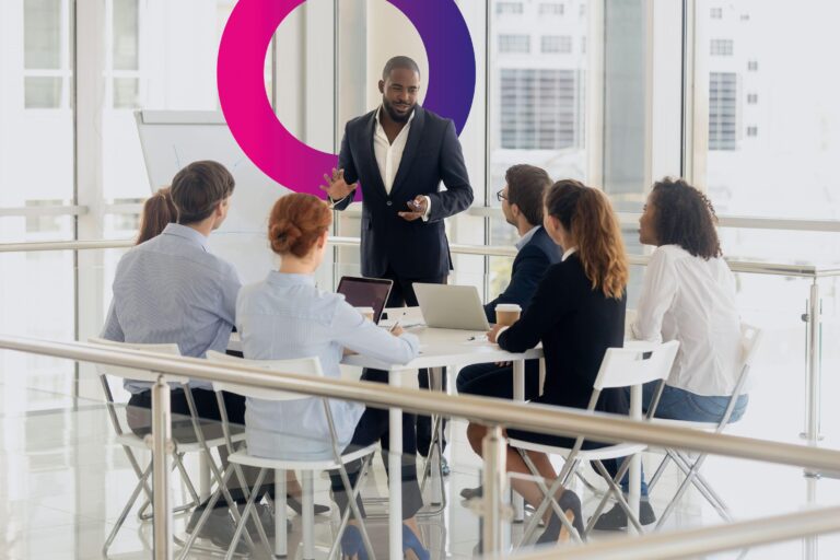 A man stands and speaks to five seated colleagues in a modern office, with laptops and coffee cups on the table. A whiteboard and a circular graphic are visible in the bright background.