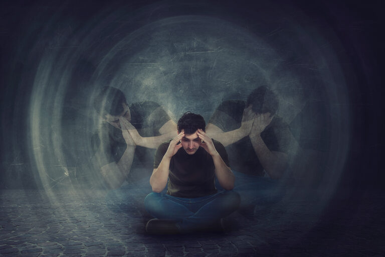 A person sits cross-legged on the ground, holding their head with both hands, as blurred duplicates of themselves clutch their face in distress, suggesting anxiety or mental struggle.