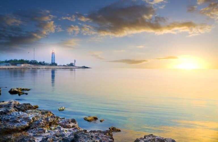Rocky shoreline with calm water at sunset, a glowing sun near the horizon, and a lighthouse visible in the distance with a partly cloudy sky.