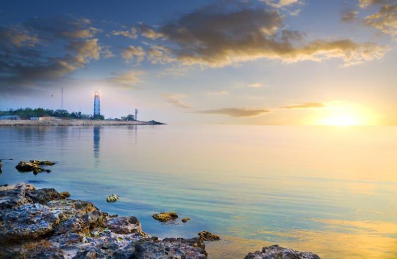 Rocky shoreline with calm water at sunset, a glowing sun near the horizon, and a lighthouse visible in the distance with a partly cloudy sky.