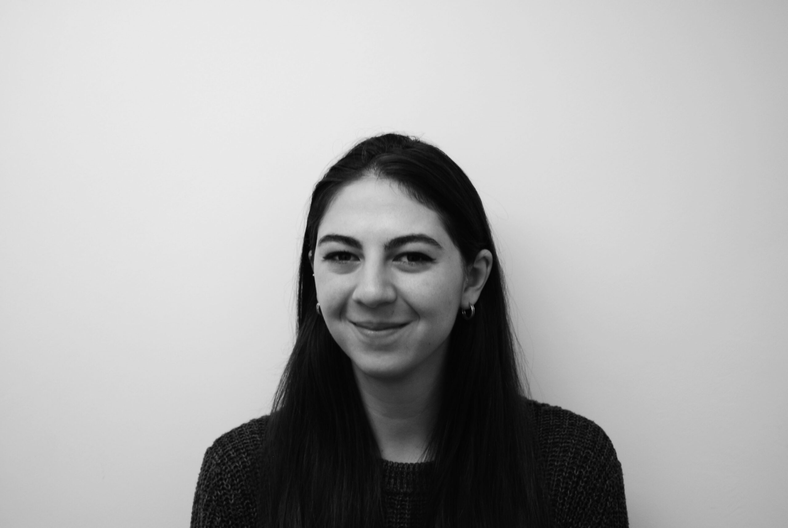 A young woman with long dark hair and hoop earrings smiles softly at the camera against a plain, light-coloured background. The photo is in black and white.