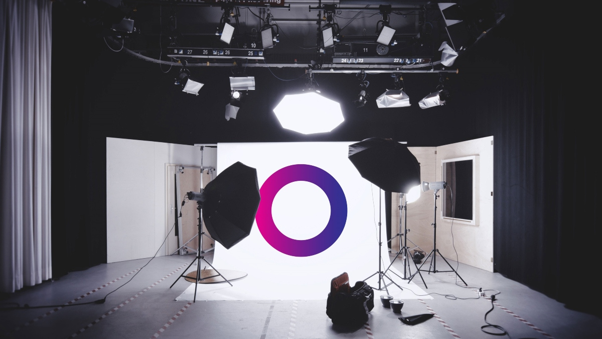 A photography studio set-up with lights, softboxes, and tripods focused on a backdrop displaying a bold purple and pink circle logo. Some photography equipment is visible on the floor.