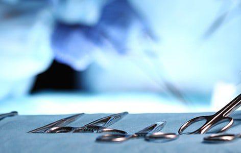 A close-up of surgical instruments, including scissors and forceps, laid out on a sterile table, with a blurred figure in surgical theatre scrubs visible in the background.