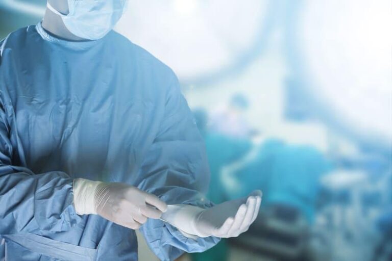 A surgeon wearing a mask and gloves adjusts his glove in an operating theatre, with bright surgical lights and blurred medical staff visible in the background.