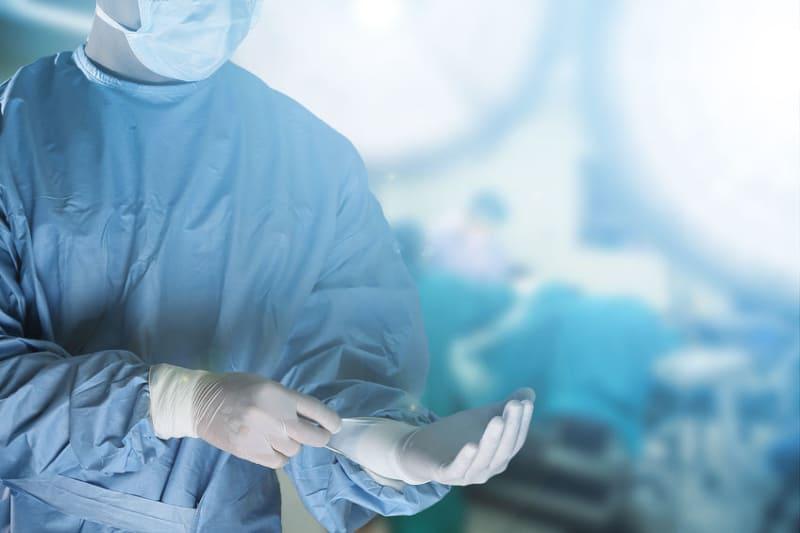 A surgeon wearing a mask and gloves adjusts his glove in an operating theatre, with bright surgical lights and blurred medical staff visible in the background.