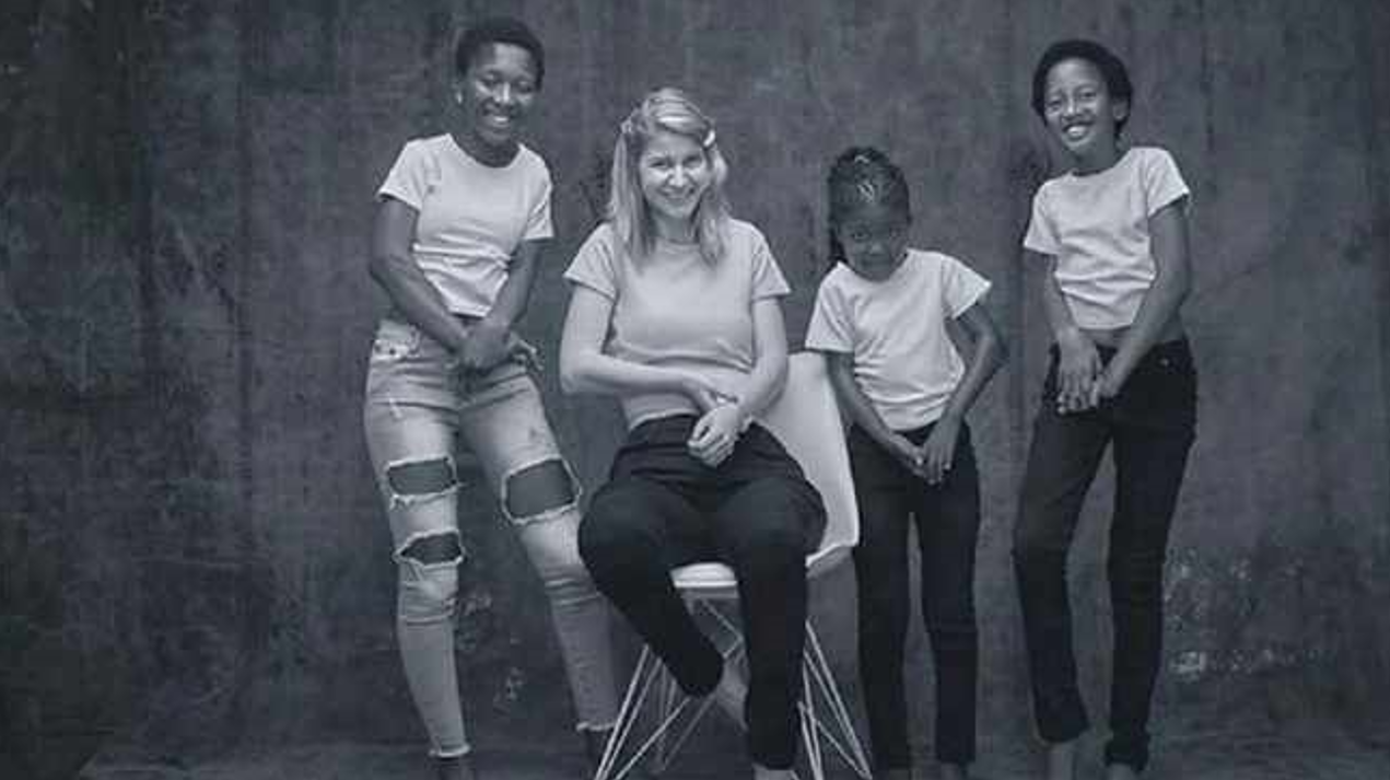 Four children pose together in a studio. Three stand, smiling, wearing T-shirts and jeans; one sits in a chair in the centre. The background is plain and the image is in black and white.