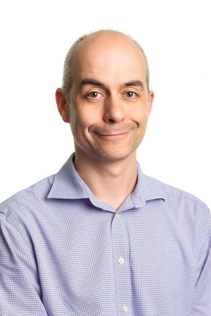 A man with a receding hairline, wearing a light purple checked shirt, smiles slightly whilst facing the camera against a plain white background.