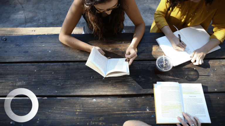 Three people sit at a wooden picnic table reading books and taking notes. A clear plastic cup with a drink and a notebook are on the table. Sunlight casts shadows across the scene.