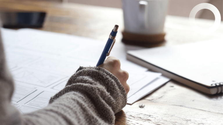 A person in a grey jumper writes on papers at a wooden table with a pen. A notebook, mobile phone, and white mug are nearby.
