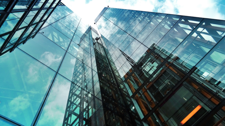 Looking up at modern glass skyscrapers reflecting clouds and blue sky, with steel beams and geometric patterns visible in the architecture.
