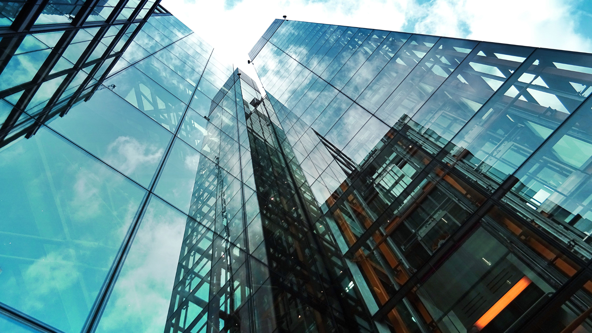 Looking up at modern glass skyscrapers reflecting clouds and blue sky, with steel beams and geometric patterns visible in the architecture.