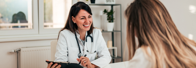 A smiling female doctor with a stethoscope talks to a patient across a desk in a bright, modern surgery.