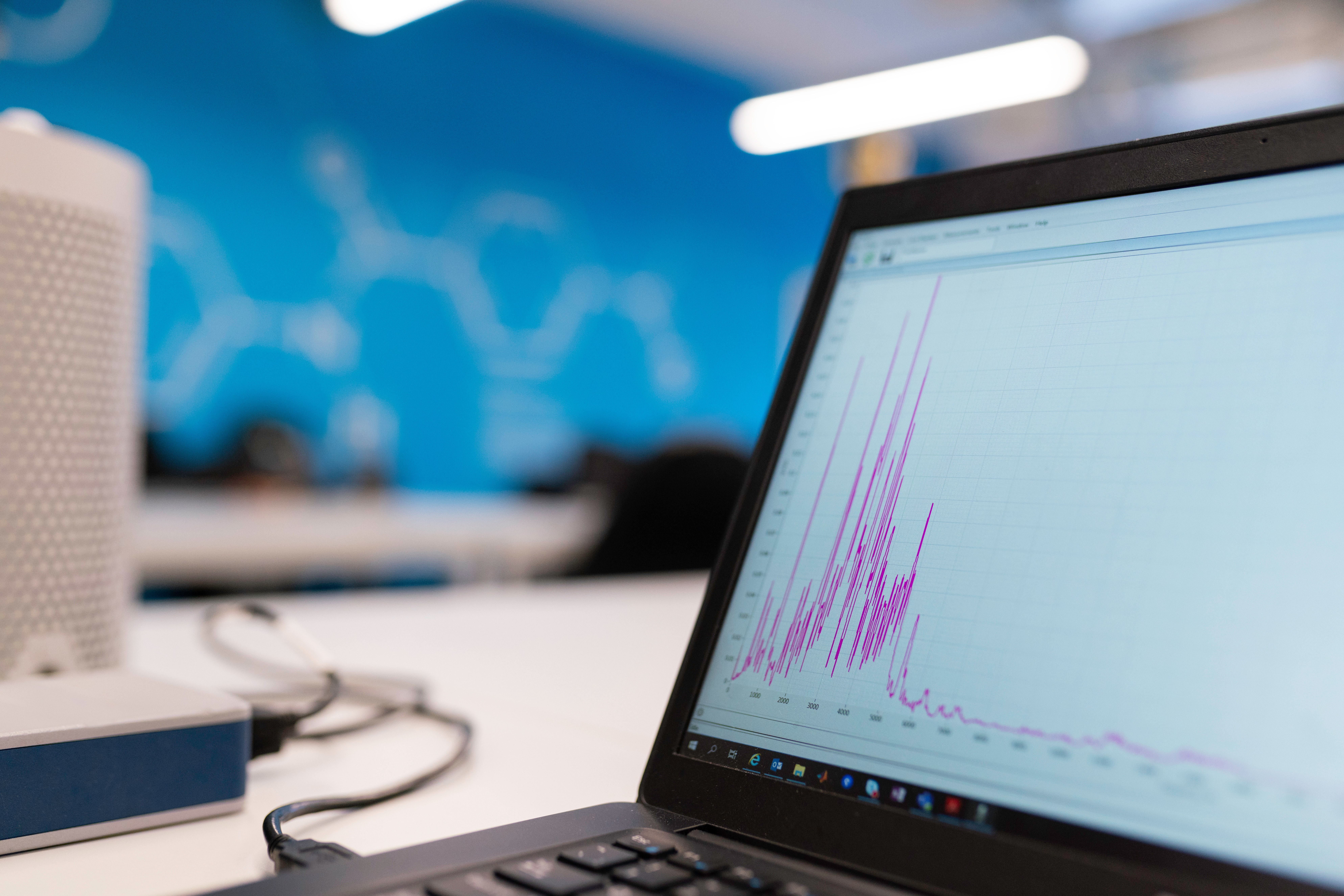 A laptop displaying a line graph with purple spikes on its screen sits on a white desk in a modern office with a blue wall in the background.