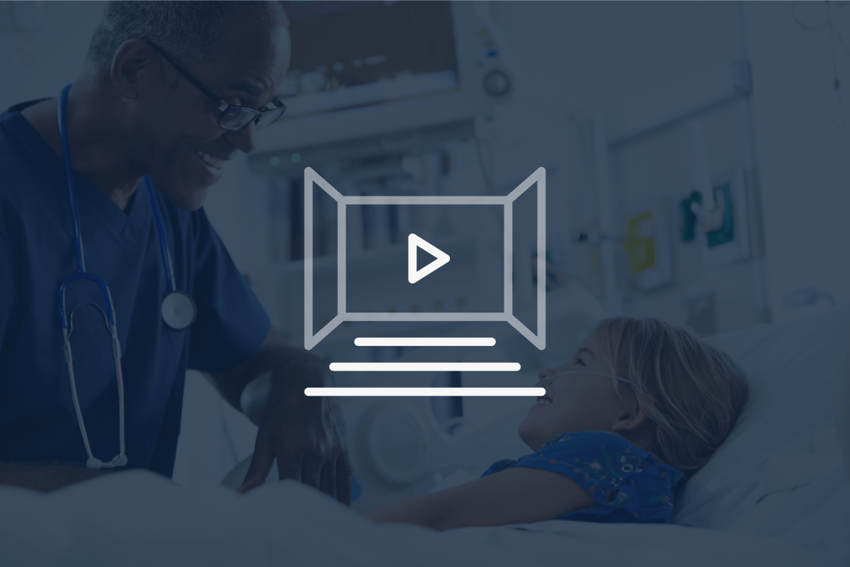 A doctor smiles and talks to a young patient lying in a hospital bed. A white play button icon overlays the image, suggesting a video.