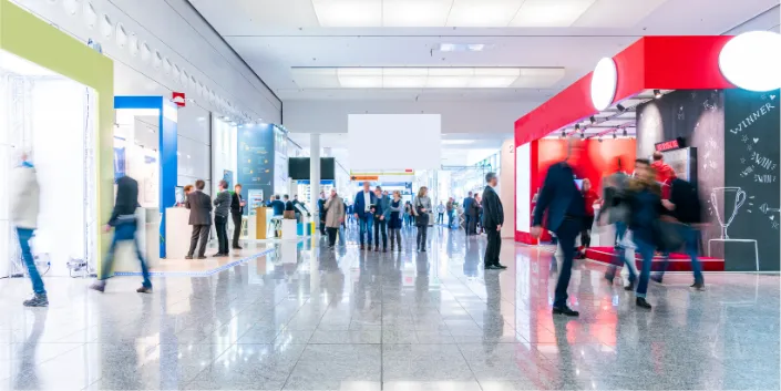 A bright, modern trade show or exhibition hall with people walking and visiting various colourful stands; the floor is shiny and reflective.