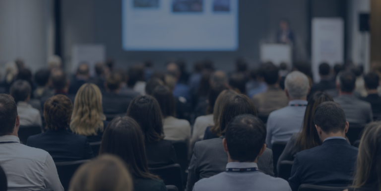 A large audience sits in rows facing a stage where a speaker presents in front of a screen displaying slides at a conference or seminar.