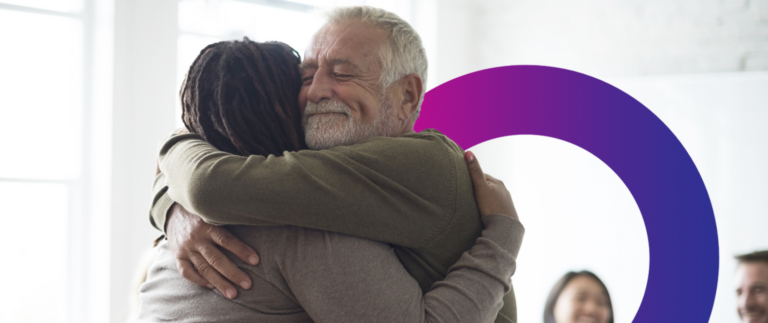 An older man and another person hug each other warmly in a bright room, with smiling people blurred in the background and a purple circle design partially visible.