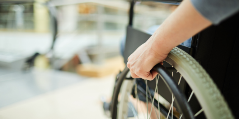 Close-up of a person's hand gripping the wheel of a wheelchair, with a blurred indoor background.