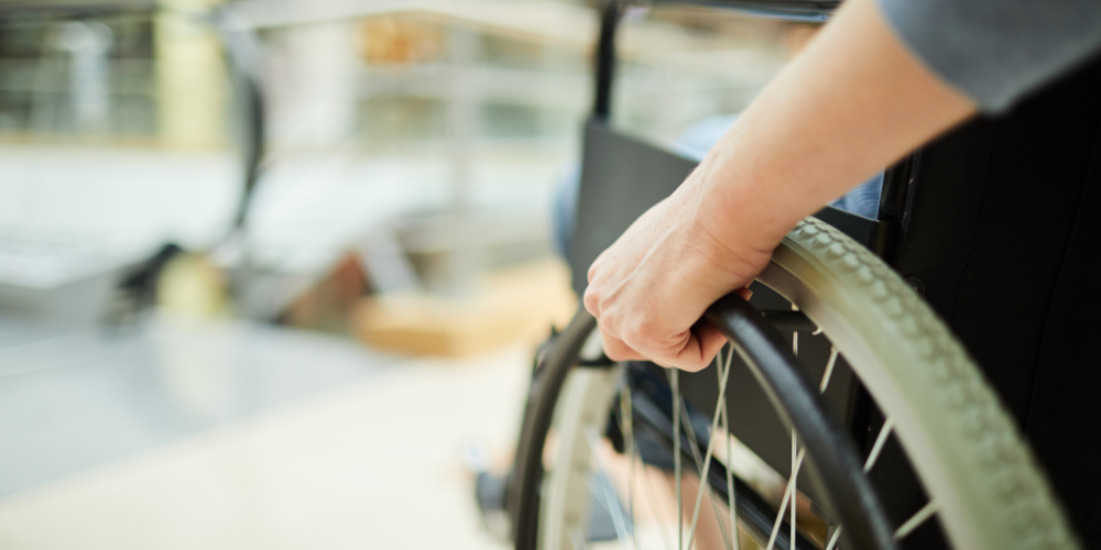 Close-up of a person's hand gripping the wheel of a wheelchair, with a blurred indoor background.
