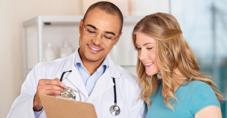 A doctor in a white coat shows a clipboard to a smiling female patient. They are standing indoors, appearing to discuss something positive.