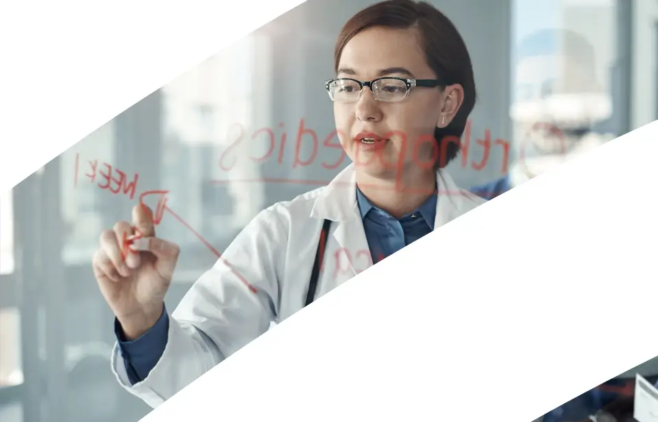 A doctor in a white coat writes notes with a red marker on a transparent board. She appears focused, with medical text and arrows visible. The image is partially covered by white diagonal shapes.