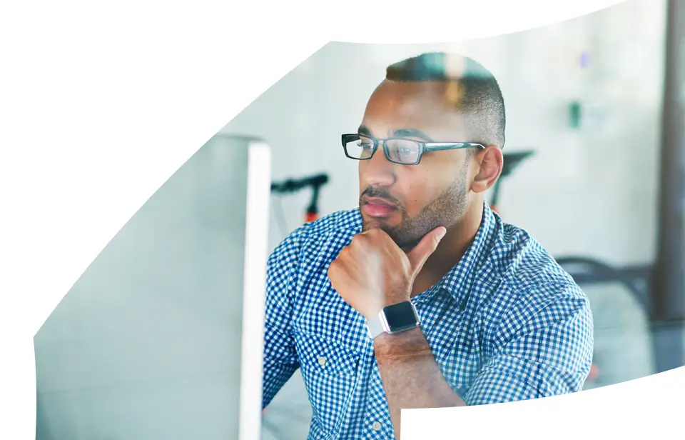 Man wearing glasses and a smartwatch sits at a desk, looking thoughtfully at a computer screen. He rests his chin on his hand and wears a blue chequered shirt. The background is blurred.