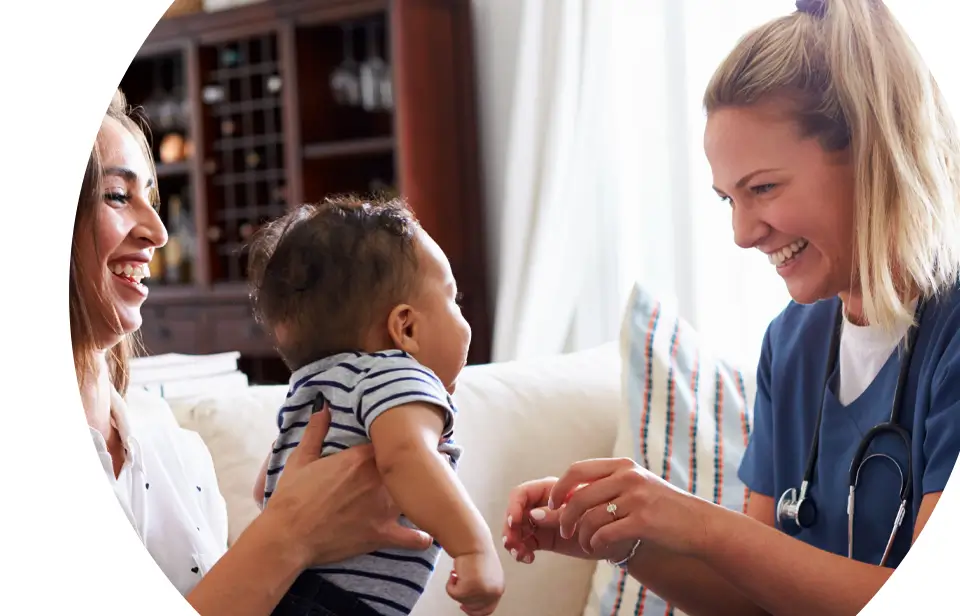 A nurse smiles and interacts with a baby held by a smiling woman, likely the mother, in a brightly lit sitting room.