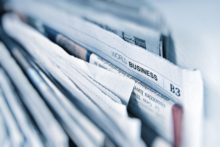 A close-up of a stack of newspapers, with one showing the World Business section in focus. The pages appear slightly fanned out and the overall tone of the image is cool and blue.