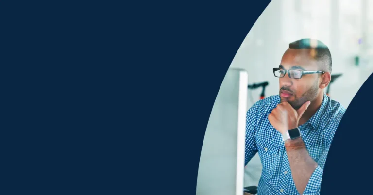 Man wearing glasses and a chequered shirt sits at a desk, looking thoughtfully at a computer screen, with a blue background and curved design on the left.
