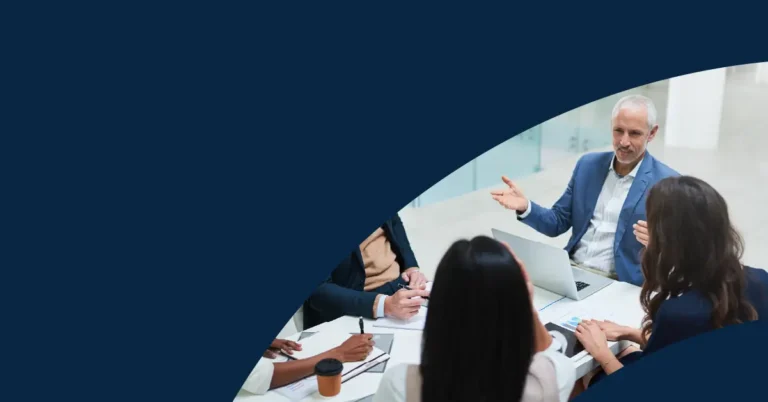 Four people sit at a meeting table with papers, a coffee cup, and a laptop. One man gestures whilst speaking. Only part of the group is visible, with a dark blue curved overlay covering most of the image.
