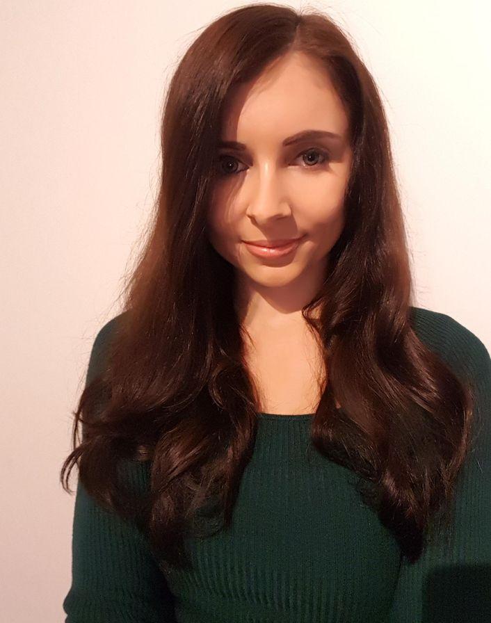 A woman with long, wavy brown hair wearing a dark green jumper is standing against a plain white background, smiling gently at the camera.
