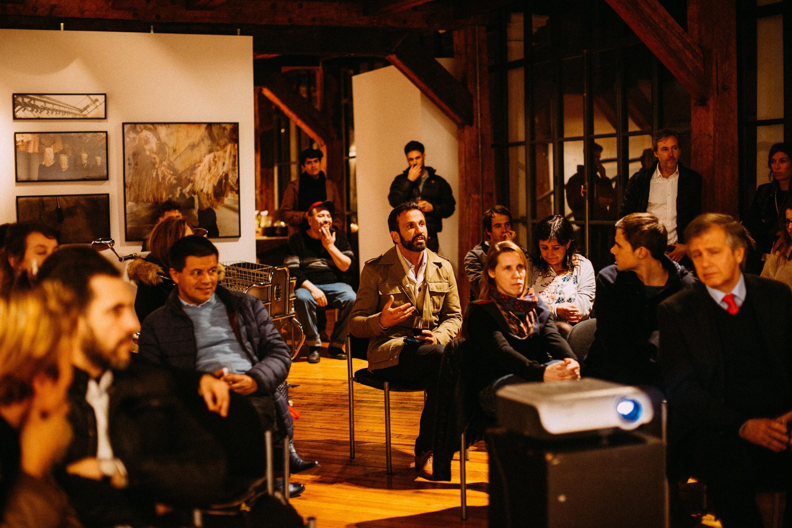 People sit in a warmly lit room, attentively listening to a speaker. Some look engaged, one man gestures whilst speaking, and a projector is visible in the foreground. Artwork hangs on the walls.