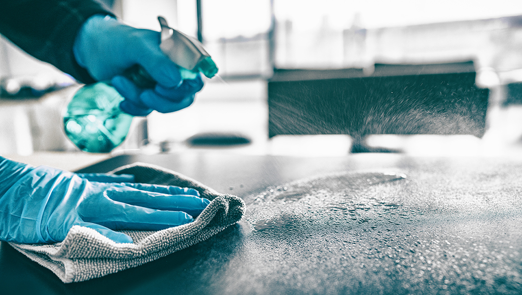 A person wearing blue gloves sprays cleaner onto a surface and wipes it with a cloth, cleaning and disinfecting a worktop.