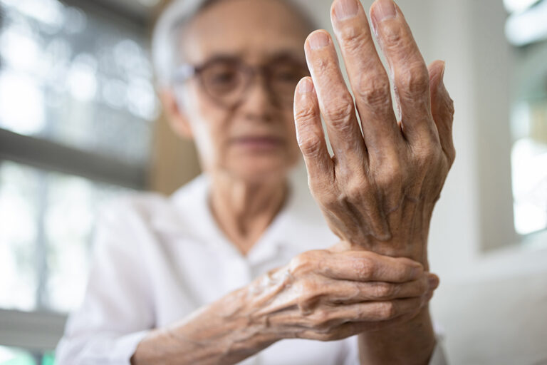 An older person holds their wrist with one hand, looking at raised fingers, possibly indicating hand pain or discomfort.