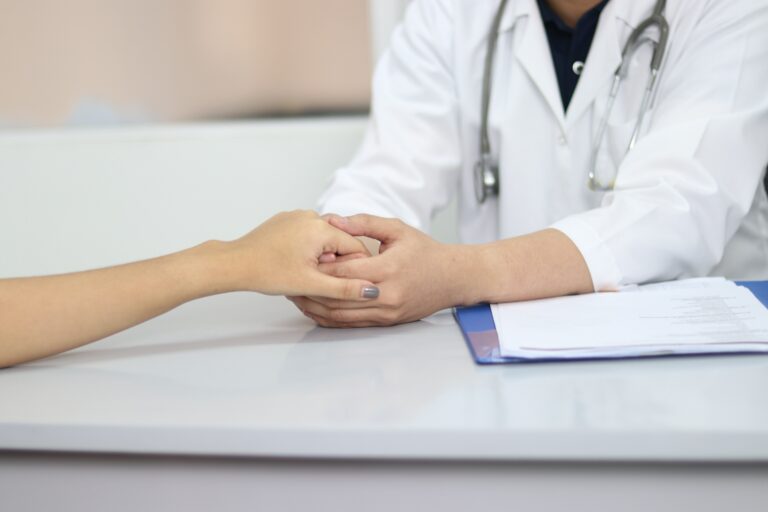A doctor in a white coat gently holds a patient's hand across a desk, offering comfort and support. A clipboard with paperwork rests on the desk beside them.