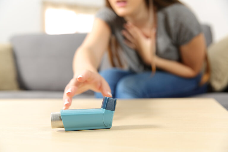 A woman sitting on a sofa clutches her chest and reaches for an inhaler on a table, suggesting she may be experiencing breathing difficulties.