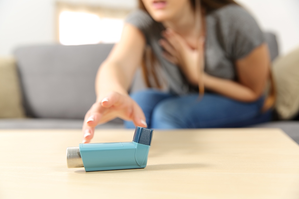 A woman sitting on a sofa clutches her chest and reaches for an inhaler on a table, suggesting she may be experiencing breathing difficulties.