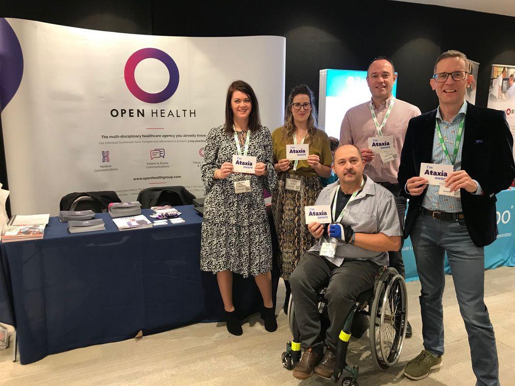 Five people, one in a wheelchair, stand together at an Open Health event stand, each holding a card that says Ataxia. There are brochures and banners visible on the table and in the background.