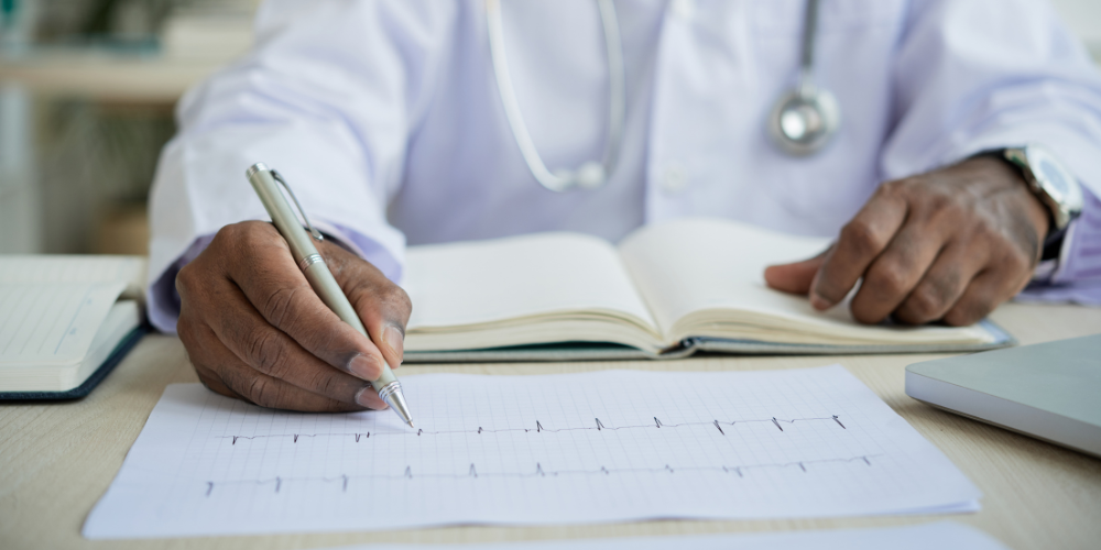 A doctor wearing a white coat reviews and writes notes on an electrocardiogram (ECG) chart, with an open notebook and a stethoscope in the background.