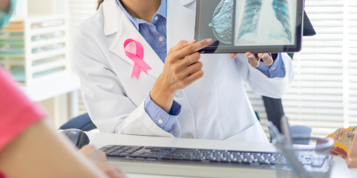 A doctor wearing a pink ribbon points to a lung X-ray on a computer screen whilst discussing it with a patient across the desk.