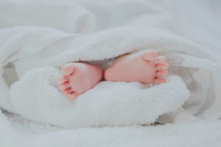 A close-up of a baby's feet peeking out from under a soft white blanket, with the toes slightly curled and surrounded by fluffy fabric.