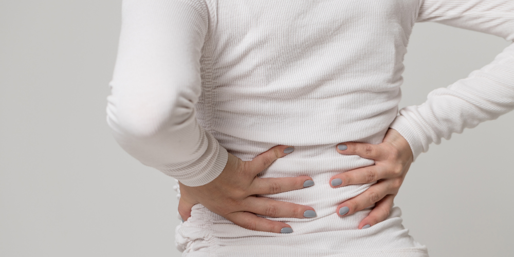 A person in a white shirt holds their lower back with both hands, suggesting discomfort or back pain.