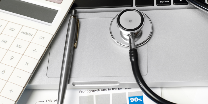 A stethoscope rests on a laptop keyboard beside a pen and a calculator, with financial documents partially visible underneath.