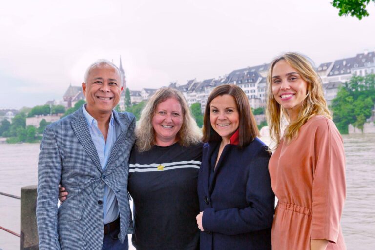 Four people stand together smiling by a river, with historic buildings and greenery in the background. They appear to be enjoying a pleasant day outdoors.