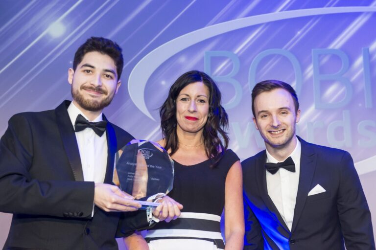 Three people in formal attire stand together at an awards event; the man on the left holds a glass award whilst all three smile at the camera, with a blue-lit BOBI Awards backdrop behind them.