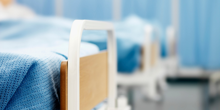 A close-up of a hospital bed with light blue sheets and a wooden headboard, set in a row in a medical facility. The background is blurred, showing more beds and blue privacy curtains.