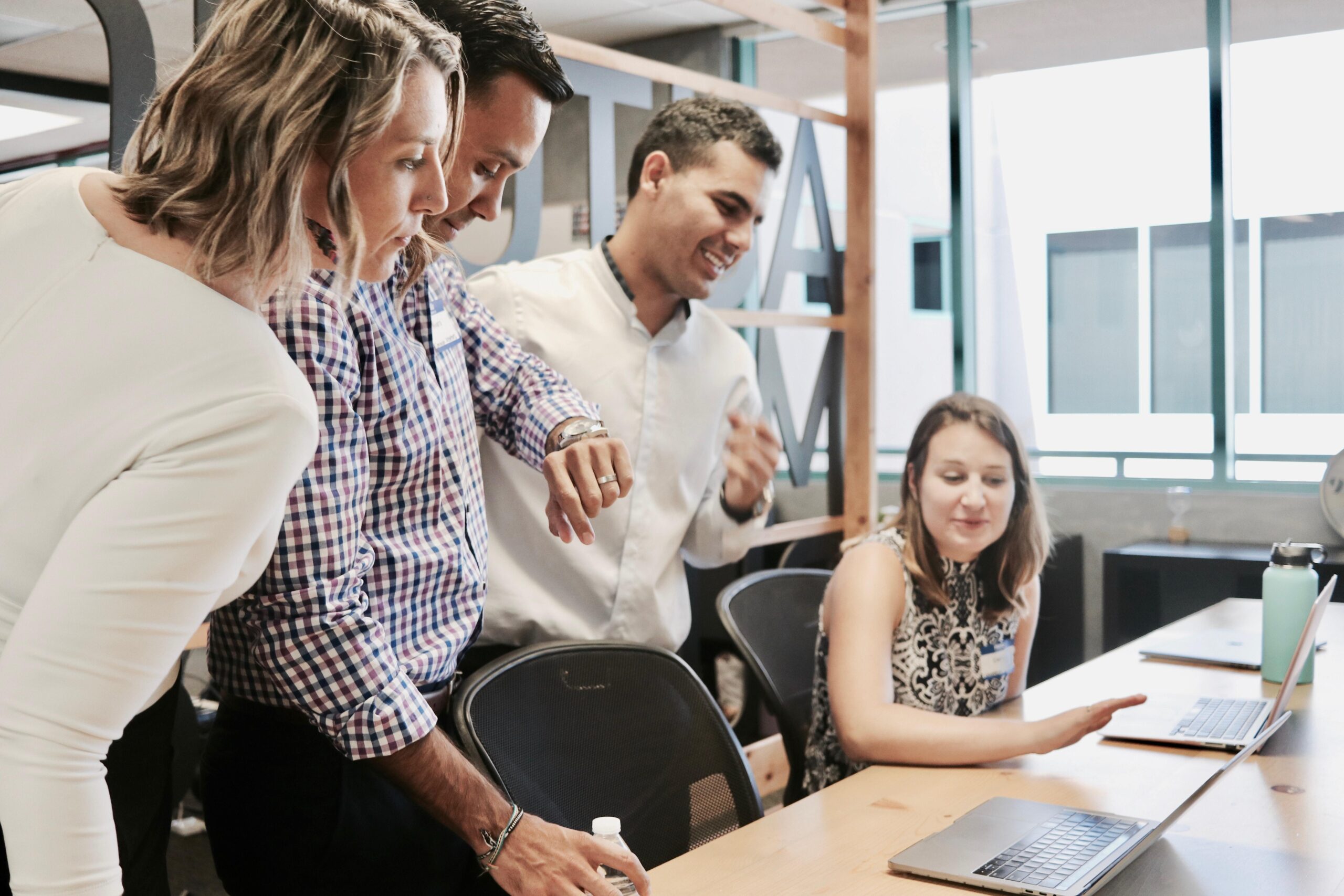 Four people gather round a table in an office, looking at a laptop screen and smiling. One person sits at the laptop, while the others stand, appearing engaged in a discussion or presentation.