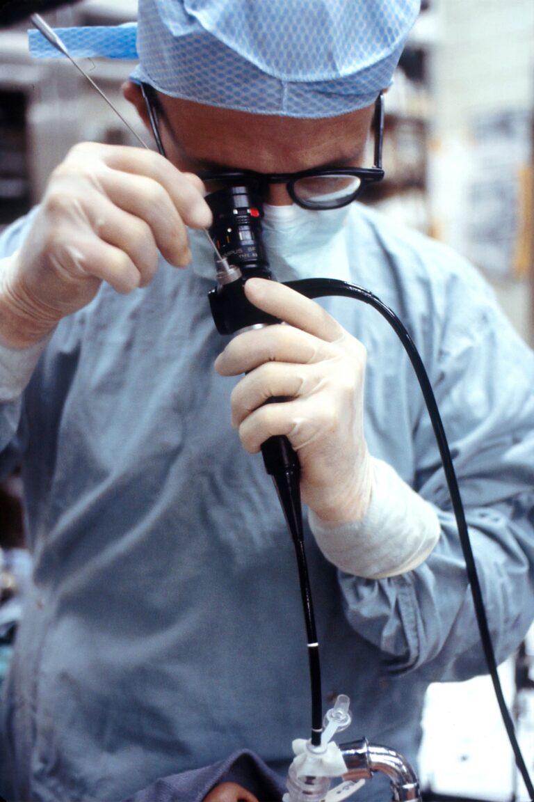 A medical professional wearing scrubs, gloves, and a mask looks into an endoscope during a procedure in a clinical setting.