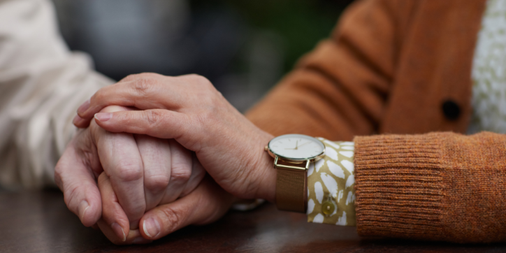 Close-up of two people holding hands; one person wears a brown jumper, patterned sleeve, and a wristwatch. The gesture suggests comfort, support, or affection between them. The background is softly blurred.